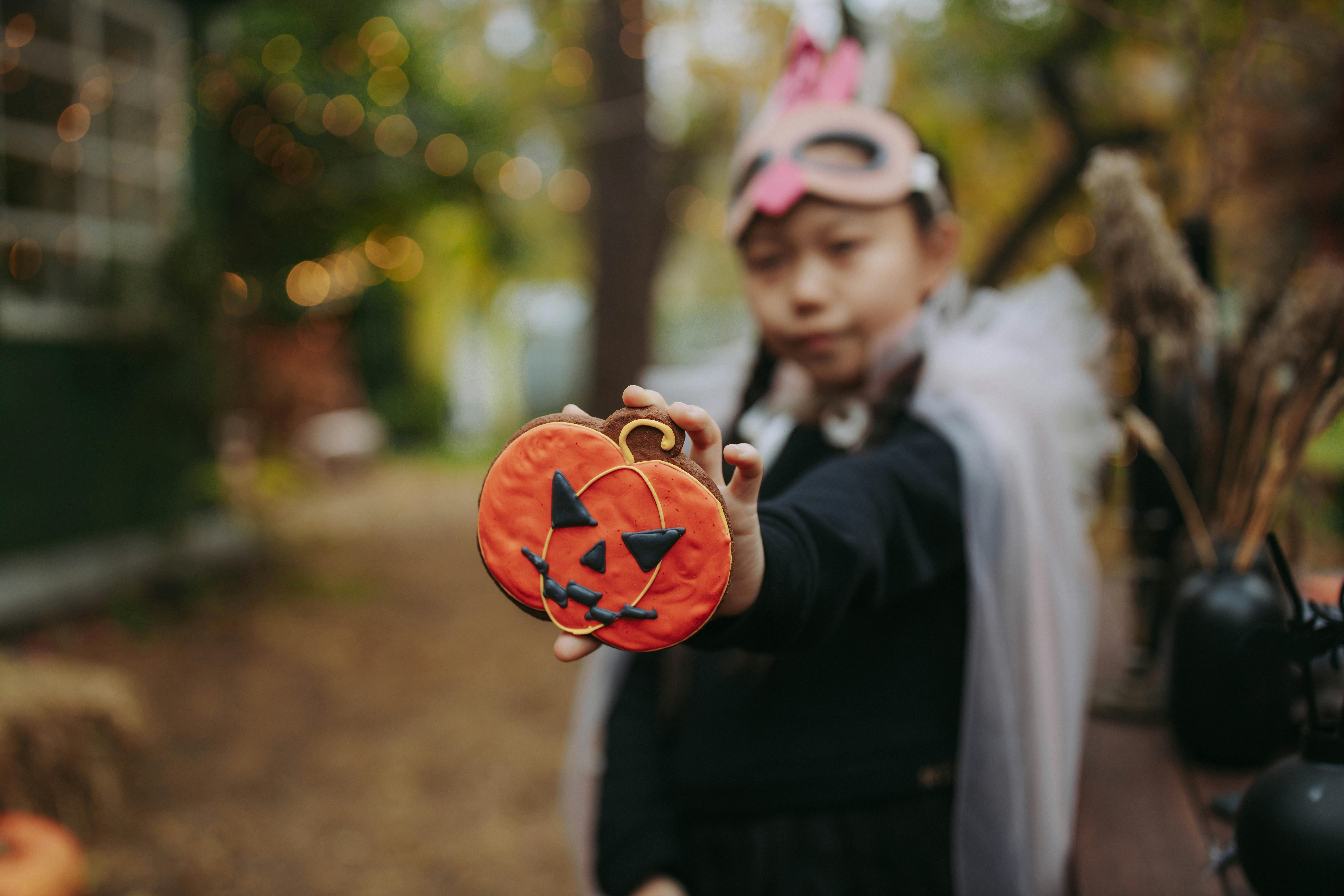 Girl in Halloween mask and cape showing pumpkin cookie to camera
